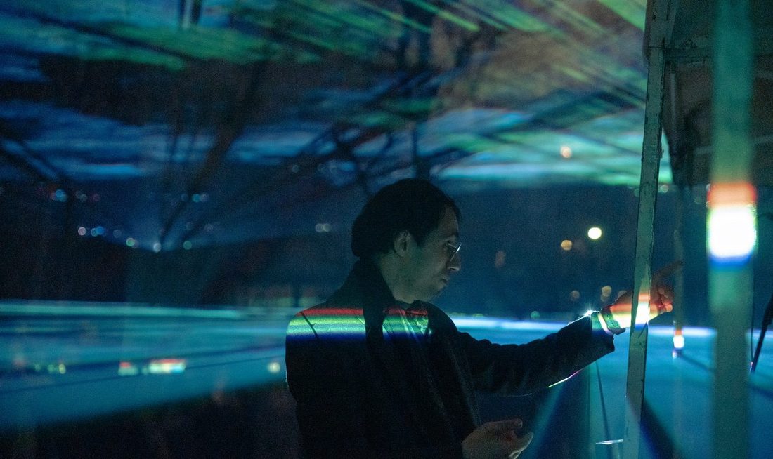 A visitor tracing a lit panel, spectral light across the ceiling